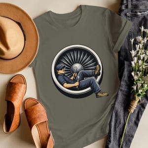 May include: Olive green t-shirt featuring a cartoon illustration of a mechanic relaxing inside a jet engine. The mechanic is wearing a blue uniform and holding tools. The shirt is paired with brown sandals, a brown hat, and blue jeans.