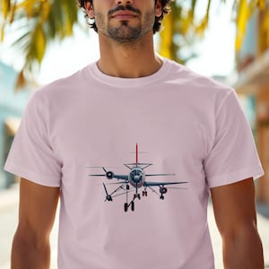 May include: A light pink t-shirt featuring a detailed illustration of a vintage airplane. The aircraft is depicted in shades of gray, blue, and red, with visible propellers and landing gear. The design is centered on the shirt.