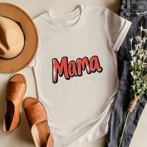 May include: A white t-shirt with the word "Mama" in red, outlined in black. The shirt is displayed with brown sandals, a brown hat, and dark blue jeans. A bouquet of white flowers is also present.