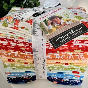 May include: A stack of colorful fabric squares with a white measuring tape and a tag that reads "Fruit Cocktail Fig Tree & Co" and "moda Fabrics + Supplies".