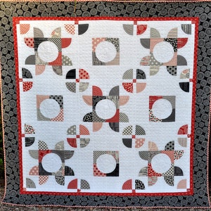 May include: A white quilt with a geometric pattern of red, black, and white circles and half circles. The quilt is framed with a black and white border.