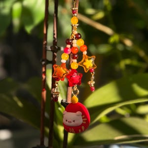 May include: A decorative charm featuring a gold star, colorful beads, and star-shaped accents. The charm includes a red, egg-shaped pendant with a pig design. The charm is suspended from a gold ring.