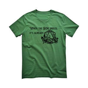 Puede incluir: Una camiseta verde de manga corta con un gráfico negro de un dado de veinte caras, una calavera y una cinta con el texto "TOO LATE". El texto sobre el dado dice "WHEN THE DM SMILES, IT'S ALREADY". El diseño está centrado en el pecho.