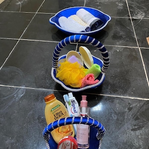 Peut inclure: Deux paniers en tissu bleu avec bordure et anses argentées, l'un contenant une fleur de douche jaune, un savon violet et deux petits tissus, l'autre une bouteille Garnier Ultra Doux, du dentifrice Signal, une brosse à dents et un pot rouge.