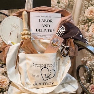 May include: A cream-colored tote bag with the text "Prepared for the Journey" and a heart-shaped graphic containing a baby silhouette. The bag contains a wooden comb, a wooden massage tool, and a card that says "Welcome to Labor and Delivery".