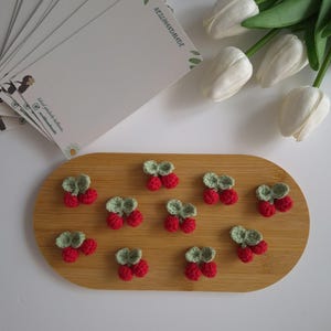 Puede incluir: Cerezas de ganchillo hechas a mano con hojas verdes, dispuestas sobre una bandeja ovalada de madera. Las cerezas son de color rojo brillante. Varias tarjetas blancas con el texto "NESLIHANDMADE" están en el fondo, junto con tulipanes blancos.