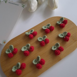 Peut inclure: Des cerises crochetées à la main, avec des feuilles vertes, disposées sur une planche en bois. Les cerises sont d'un rouge vif, et les feuilles sont vert clair. La planche est marron clair, avec des tulipes blanches et un bloc-notes en arrière-plan.
