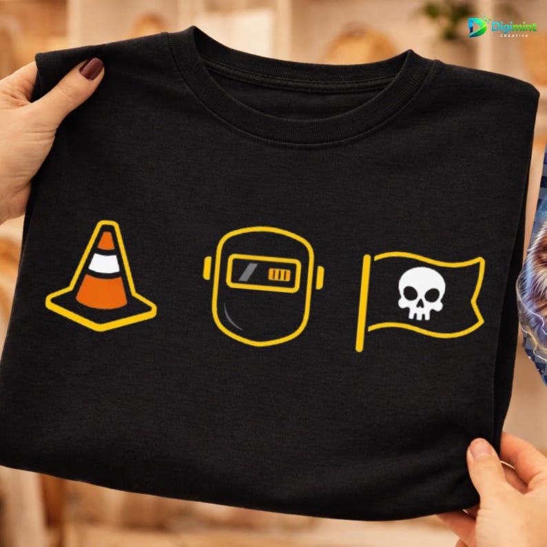Op de afbeelding: Zwart T-shirt met drie geel omrande afbeeldingen: een verkeerskegel, een lasmasker en een piratenvlag met een schedel. De afbeeldingen zijn gecentreerd op het shirt. Het shirt wordt door handen vastgehouden.