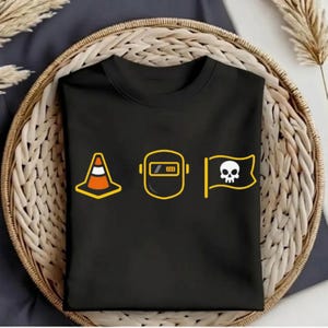 Op de afbeelding: Zwart t-shirt met drie gele en witte afbeeldingen: een verkeerskegel, een lasmasker en een vlag met een schedel. De afbeeldingen zijn geel omlijnd. Het t-shirt is opgevouwen en in een geweven mand geplaatst.
