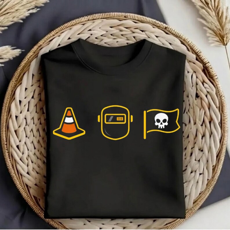 Op de afbeelding: Zwart t-shirt met drie gele en witte afbeeldingen: een verkeerskegel, een lasmasker en een vlag met een schedel. De afbeeldingen zijn geel omlijnd. Het t-shirt is opgevouwen en in een geweven mand geplaatst.