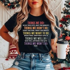 May include: Black t-shirt with colourful text including "THINGS WE DID: Built this city, shot the sheriff. THINGS WE DIDN'T DO: Start the fire, shoot the deputy." The shirt also has other phrases.