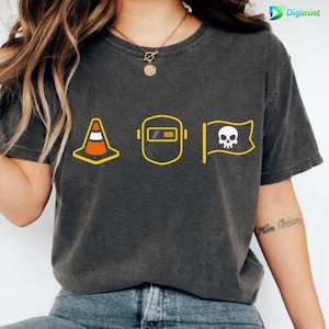Op de afbeelding: Donkergrijs t-shirt met gele en witte afbeeldingen. Het ontwerp bevat een verkeerskegel, een lasmasker met een batterij-indicator en een piratenvlag met een schedel. Het t-shirt heeft een vintage uitstraling.