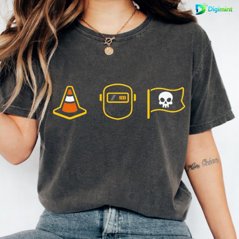 Op de afbeelding: Donkergrijs t-shirt met gele en witte afbeeldingen. Het ontwerp bevat een verkeerskegel, een lasmasker met een batterij-indicator en een piratenvlag met een schedel. Het t-shirt heeft een vintage uitstraling.