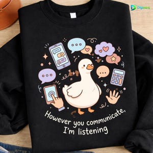 Puede incluir: Sudadera negra con una ilustración de un pato de dibujos animados rodeado de bocadillos, una calculadora y una mano. El texto "However you communicate, I'm listening" está impreso debajo.