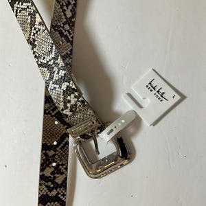 May include: A snakeskin-patterned belt with a silver-toned buckle. The belt is in shades of brown, gray, and white. A white tag with the text "Nicole Miller NEW YORK" is attached. The belt is a size L.