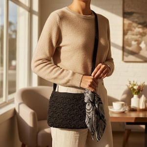May include: A black, textured crossbody bag with a matching strap and a decorative scarf. The bag is worn over the shoulder. The scarf has a black and white paisley pattern. The person is wearing a tan sweater and light-colored pants.