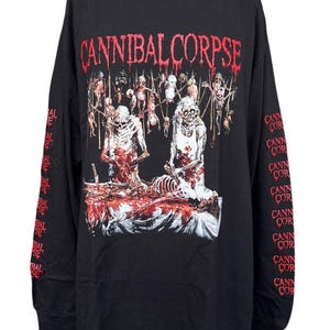 Puede incluir: Una camiseta negra de manga larga con el texto rojo CANNIBAL CORPSE en la parte superior. El gráfico muestra dos figuras esqueléticas de pie sobre una mesa con restos. El texto CANNIBAL CORPSE se repite en rojo en ambas mangas.