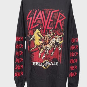 May include: A black long-sleeve t-shirt featuring a red Slayer logo at the top and a graphic of horned figures in yellow and red. The text HELL AWAITS is printed below the graphic, and the word Slayer is repeated in red down both sleeves.