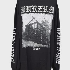 Peut inclure: Un t-shirt noir à manches longues avec une impression graphique blanche sur la poitrine et les manches. Le motif central représente une structure en bois avec le texte BURZUM au-dessus et Aske en dessous. BURZUM est imprimé sur les manches.