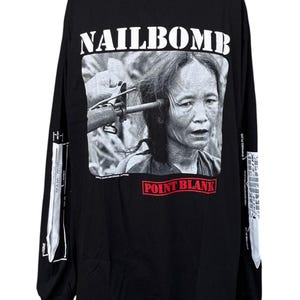 May include: A black long-sleeve t-shirt featuring a white graphic print of a person with a rifle pointed at their head. The text NAILBOMB appears in white above the image, and the words POINT BLANK are printed in red below the graphic.