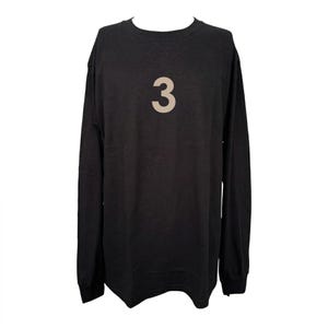 May include: Black long-sleeved top featuring the number "3" in a light tan shade on the front. The top has a crew neck and a loose fit. The sleeves are long, reaching the wrists. The garment is made from a soft fabric.