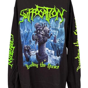 Puede incluir: Una camiseta negra de manga larga con un logotipo verde neón brillante que dice Suffocation en la parte superior. El gráfico central muestra un paisaje surrealista azul oscuro con un pilar central y una criatura. El texto Breeding the Spawn aparece debajo.