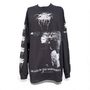 Op de afbeelding: Zwart shirt met lange mouwen met een wit bandlogo en een grijswaardenafbeelding van een persoon met lang haar. Witte afbeeldingen sieren de mouwen. De tekst "A Blaze in the Northern Sky" staat onderaan het shirt.