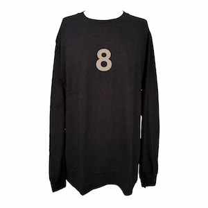 May include: A black long-sleeve shirt featuring a crew neckline and a large, centered beige numeral eight printed on the chest. The garment has a straight hemline and long sleeves with ribbed cuffs, providing a simple and uniform appearance.
