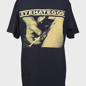 Może przedstawiać: Czarny t-shirt z krótkim rękawem z grafiką w kolorze żółtym i czarnym. Nadruk przedstawia napis EYEHATEGOD w prostokątnej ramce nad obrazem dłoni trzymającej strzykawkę przy ramieniu. Wzór jest wyśrodkowany na klatce piersiowej koszulki.