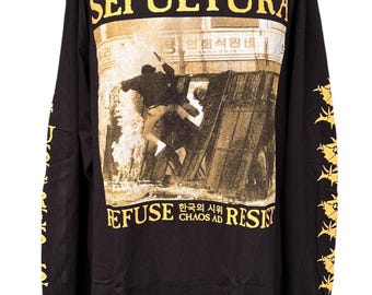 Puede incluir: Una camiseta negra de manga larga con el texto amarillo SEPULTURA en la parte superior y REFUSE CHAOS AD RESIST en la parte inferior. El gráfico central muestra una escena en blanco y negro de una persona escalando una barricada.
