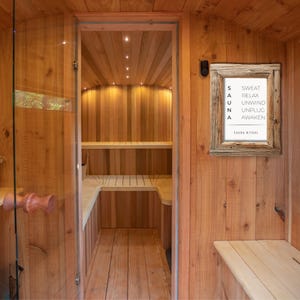May include: A wooden sauna interior with a glass door and a framed sign that reads "SAUNA" vertically, with the words "SWEAT, RELAX, UNWIND, UNPLUG, AWAKEN" below. The sauna is constructed of light-colored wood.