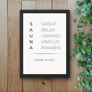May include: A framed print with the word "SAUNA" vertically stacked on the left and related words like "SWEAT" and "RELAX" to the right. The phrase "SAUNA RITUAL" is at the bottom. The frame is black, and the background is a light color.