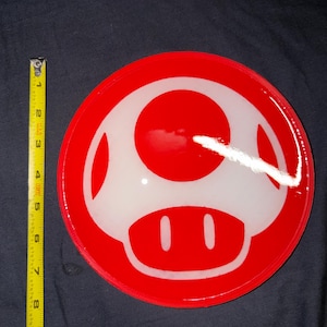 Puede incluir: Un objeto rojo y blanco con forma de hongo, probablemente un posavasos o elemento decorativo. El diseño presenta un círculo rojo con un gráfico de hongo blanco. El objeto mide aproximadamente 10 cm de diámetro.