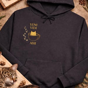 May include: Black hooded sweatshirt with a front pocket and drawstring. The design features a cat in a teacup with the words "VENI VIDI ABII" in gold lettering, along with paw prints. A cat is lying on a wooden surface.