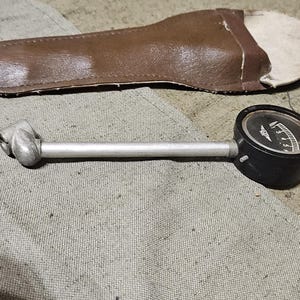 Può includere: Un manometro per la pressione dei pneumatici vintage con quadrante nero e stelo argentato, accanto a un astuccio in pelle marrone. Il manometro misura la pressione dei pneumatici. Gli oggetti sono su una superficie chiara e strutturata.
