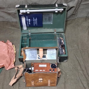 Op de afbeelding: Een militaire veldset in vintage stijl met een donkergroene metalen koffer met een zilveren staaf, een blauwe handleiding en een bruine lederen tas met een apparaat gelabeld PP-51 M met een zwarte draaiknop en een roze beschermhoes.