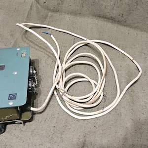 Könnte beinhalten: Ein rechteckiges Kommunikationsgerät aus salbeigrünem Metall mit der Aufschrift ACP-3, ausgestattet mit einem perforierten Lautsprechergitter, einem kleinen rechteckigen Display und einem langen weißen Stromkabel, das rechts liegt.