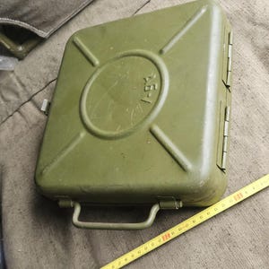 Op de afbeelding: Een olijfgroene metalen opbergkoffer met een rechthoekige vorm, een handvat aan de bovenzijde en een cirkelvormig reliëfontwerp met de tekst A5-/. Een geel meetlint ligt ernaast en toont een breedte van ongeveer 24 cm.