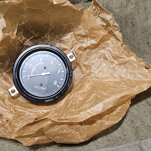 Può includere: Un manometro nero con un'ago e numeri bianchi, appoggiato su carta marrone stropicciata. Il manometro ha una lunetta nera e due linguette di montaggio. Lo sfondo è un tessuto strutturato dai toni neutri.