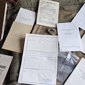 Op de afbeelding: Een verzameling van diverse papieren documenten, handleidingen en instructieboekjes met Cyrillische tekst, waaronder een bruine map met het label AP-4, diverse witte brochures en een groot technisch formulier, gerangschikt op een groene stof.