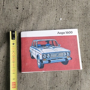 Op de afbeelding: Een rechthoekige Lada 1600 handleiding met een rode kaft voorzien van een blauw-witte illustratie van een sedan. Een geel meetlint is verticaal naast de handleiding geplaatst en toont een lengte van ongeveer 10 cm voor het boek.