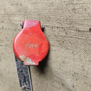 May include: A circular red metal protective cover with a hinged top, featuring the embossed text "ПУСК" on its surface. The cover is mounted on a textured olive-drab canvas material, with a small black metal identification tag hanging below it.