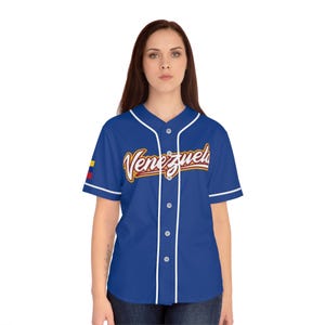 Puede incluir: Una camiseta de béisbol azul con ribetes blancos y la palabra "Venezuela" en una fuente cursiva en el pecho. La camiseta tiene mangas cortas y una parte delantera abotonada. La bandera venezolana está en la manga izquierda.