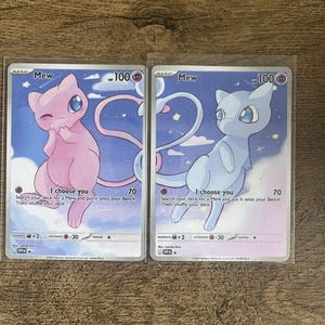 Könnte beinhalten: Zwei Pokemon-Sammelkarten mit Mew. Eine Karte zeigt ein rosa Mew, die andere ein blaues Mew, beide vor einem sternenklaren Hintergrund. Jede Karte hat den Text "I choose you" und Spieldetails.