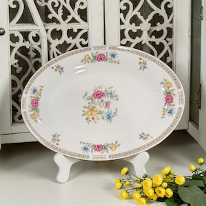 May include: Oval serving platter with a floral design. The white porcelain platter features a border with a gold trim and a pattern of pink, yellow, and blue flowers. The platter is displayed on a white stand.
