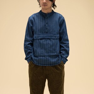 May include: A blue striped long-sleeve shirt with a mandarin collar and a chest pocket. The shirt is paired with brown corduroy pants. The outfit has a casual, yet stylish aesthetic.