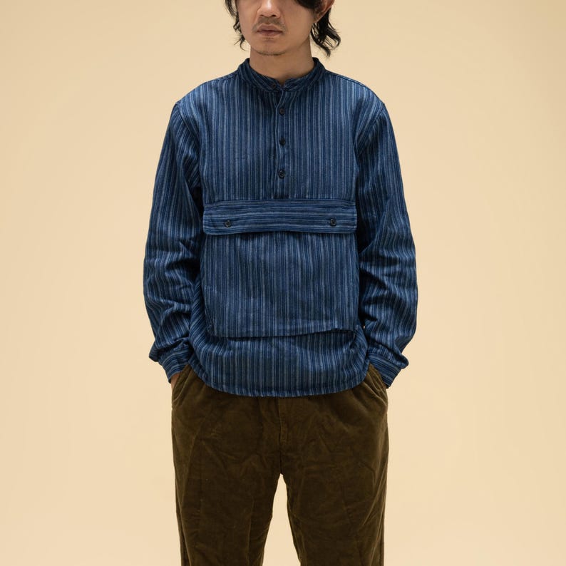 May include: A blue striped long-sleeve shirt with a mandarin collar and a chest pocket. The shirt is paired with brown corduroy pants. The outfit has a casual, yet stylish aesthetic.