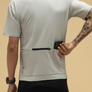 Può includere: Una maglietta sportiva grigio chiaro con una tasca con cerniera sul retro. La maglietta ha maniche corte e viene indossata con pantaloncini sportivi neri. Uno smartphone nero è nella tasca.