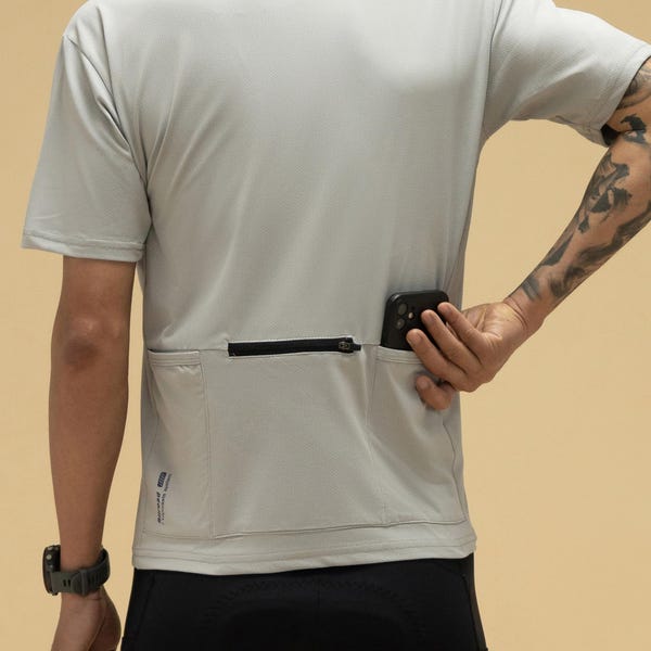 May include: A pale grey athletic t-shirt with a zipped pocket on the back. The shirt has short sleeves and is worn with black athletic shorts. A black smartphone is in the pocket.