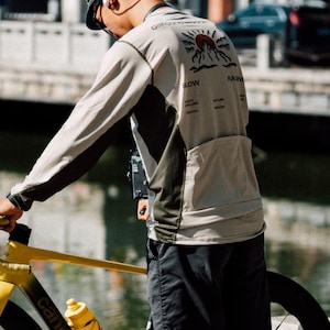 May include: A long-sleeved cycling jersey in beige and olive green, featuring a mountain and sun graphic on the back. The jersey has a pocket and the words "SLOW" and "RAZIN" printed on it. The person is holding a yellow bicycle.